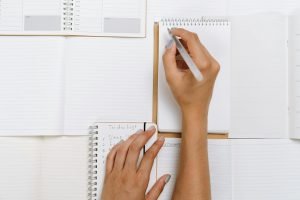 Stock photo of a pair of hands resting on top of multiple open notebooks, while one hand is writing in one of said notebooks.